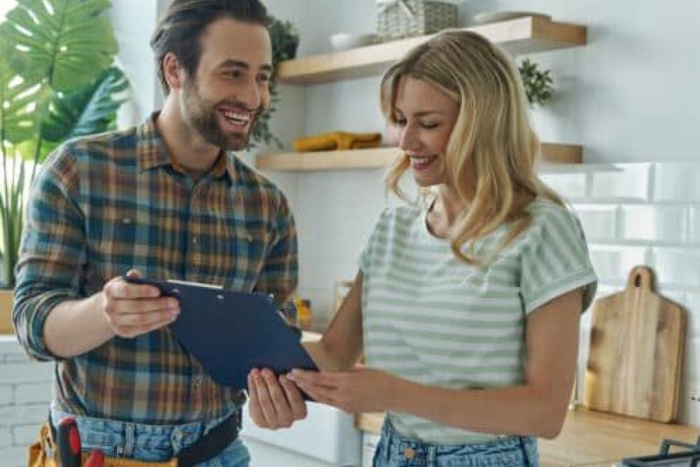 Happy couple reviewing renovation plans in a modern kitchen with white tiles and wooden shelves.