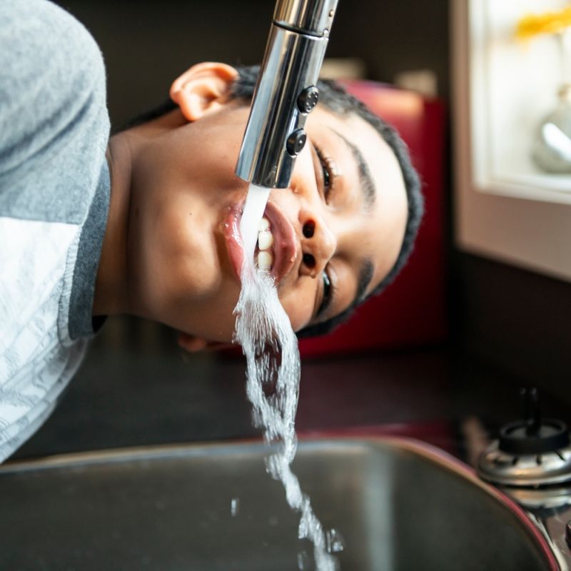 Boy drinking water directly from a kitchen faucet in a bright room.
