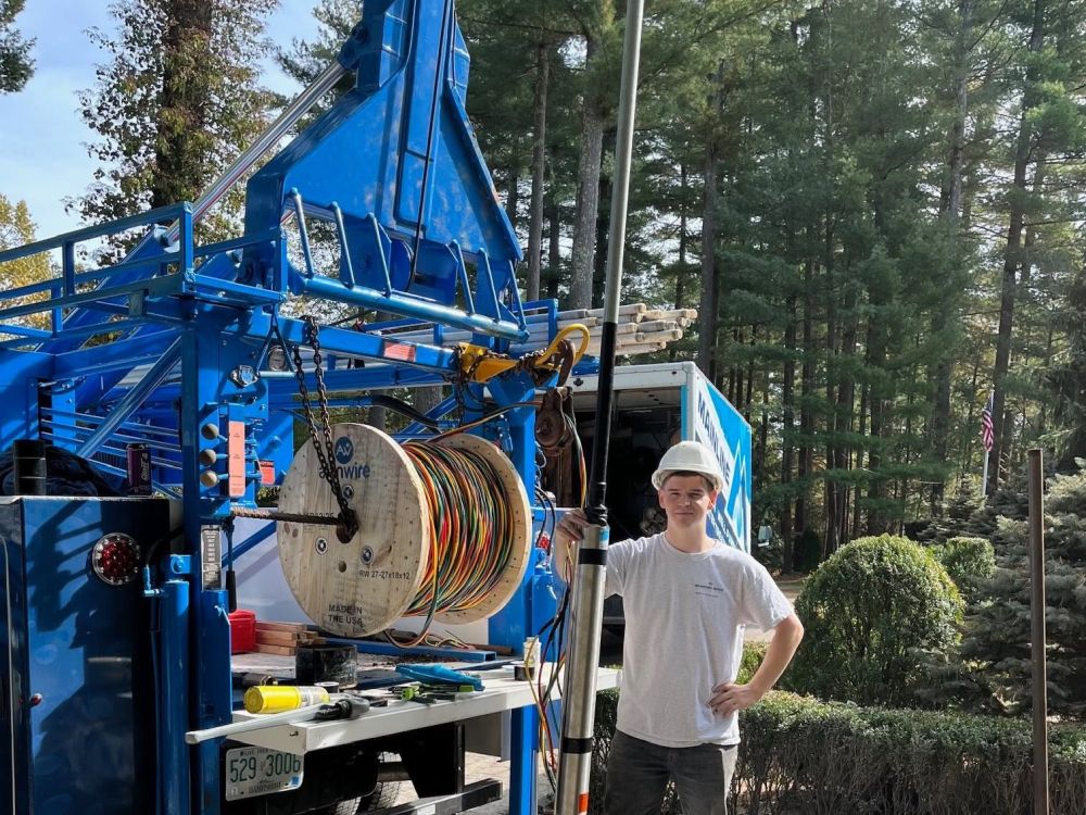 Technician in hard hat stands by a blue cable truck in a forest setting, handling electrical equipment.