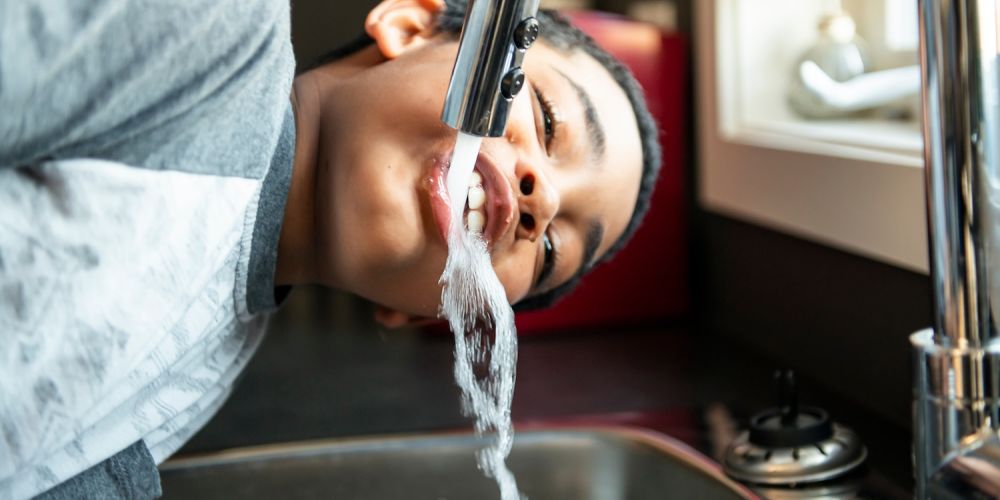 Boy drinking water directly from a kitchen faucet in a bright room.