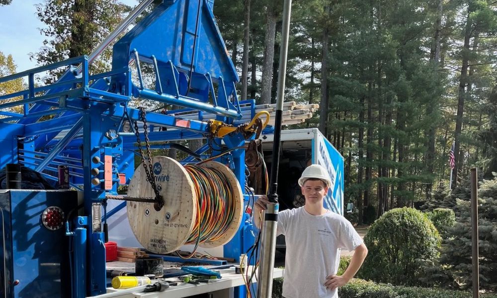 Technician in hard hat stands by a blue cable truck in a forest setting, handling electrical equipment.