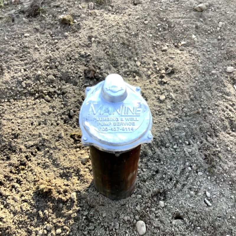 Metal well pump cover labeled Mainline Plumbing & Well on a dirt surface, providing water services information.