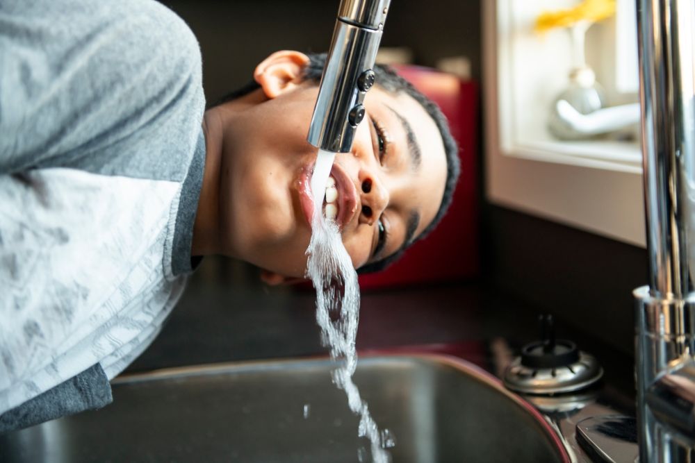 Boy drinking water directly from a kitchen faucet in a bright room.