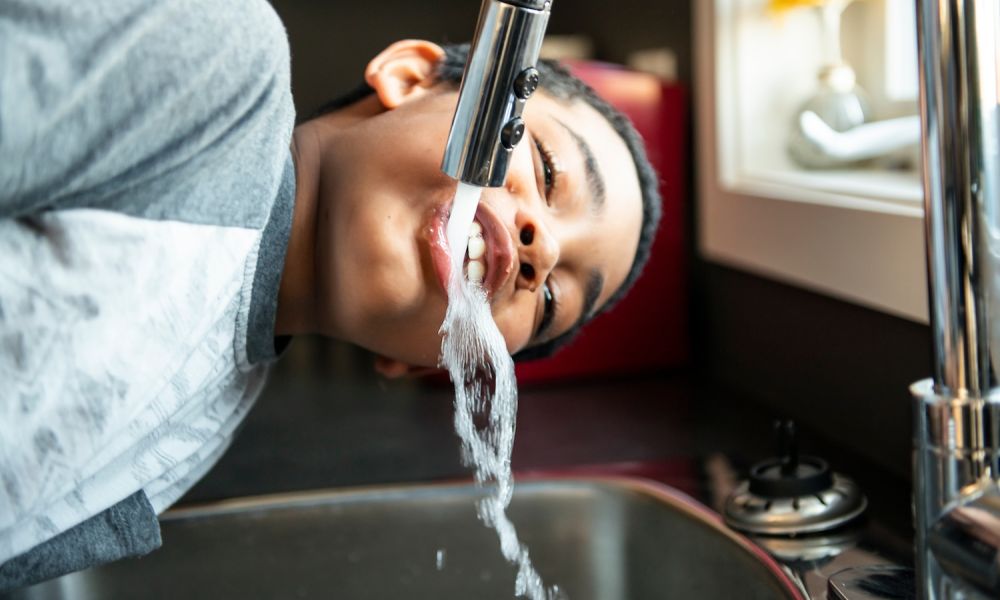 Boy drinking water directly from a kitchen faucet in a bright room.
