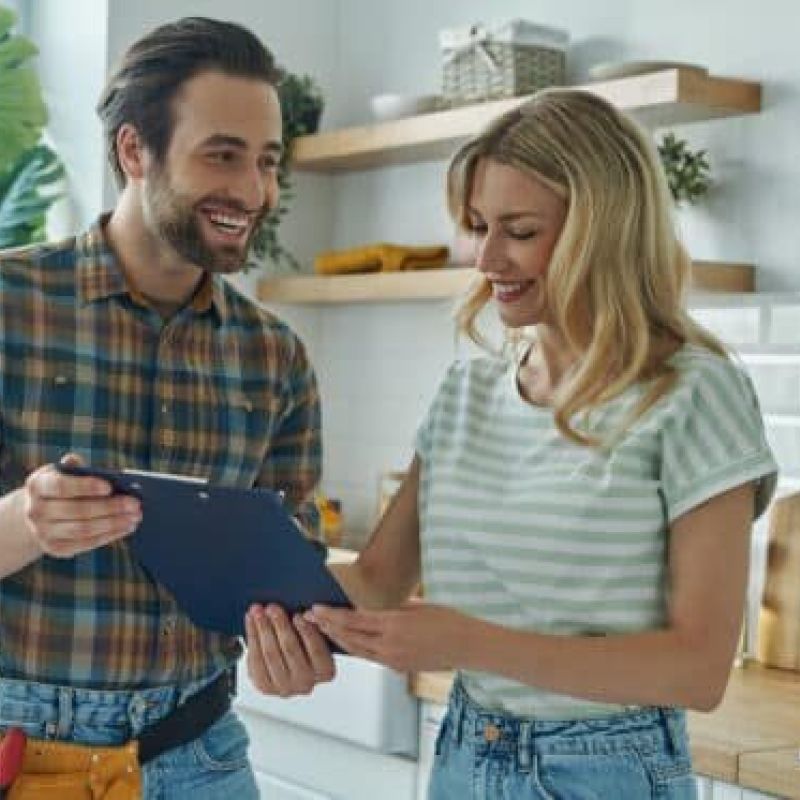 Happy couple reviewing renovation plans in a modern kitchen with white tiles and wooden shelves.